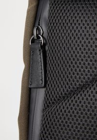 Un tissu en maille sombre et un tissu vert olive créent une surface texturée sur un sac, agrémenté de quincaillerie noire et d'une languette en cuir.