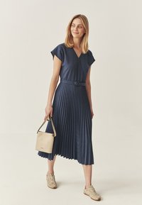Kleid in Marineblau mit Falten, kurzen Kapuffärmeln, tailliertem Gürtel, kombiniert mit einer hellbeigen Handtasche und braunen Sneakers. Glatte Stoffstruktur.