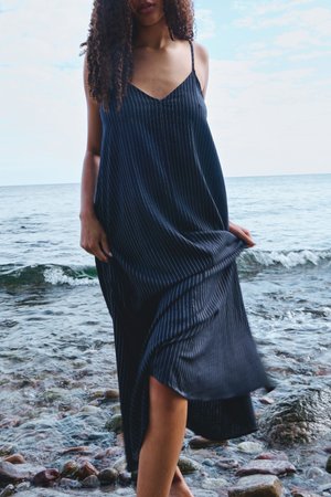 Göndör hajú nő fekete, tűszalas ujjatlan ruhában, mezítláb áll a sziklás tengerparton.