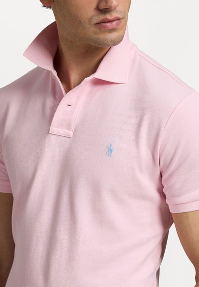 Polo roz deschis cu guler clasic, mâneci scurte și un logo brodat albastru pe piept. Material texturat cu un model subtil.