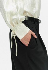 Witte satijnen blouse met een geplooide mouw en gestrikte mouwen, gecombineerd met zwarte broek. Het gladde textuur en de elegante ontwerpelementen zijn zichtbaar.