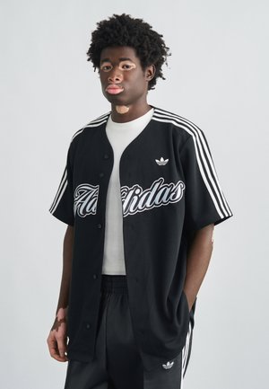 Μαύρο φαρδύ τζέρσεϊ με κουμπιά, με λευκή επιγραφή "Adidas" και τρεις λωρίδες στα μανίκια. Φορεμένο πάνω από λευκό πουκάμισο και μαύρο παντελόνι.