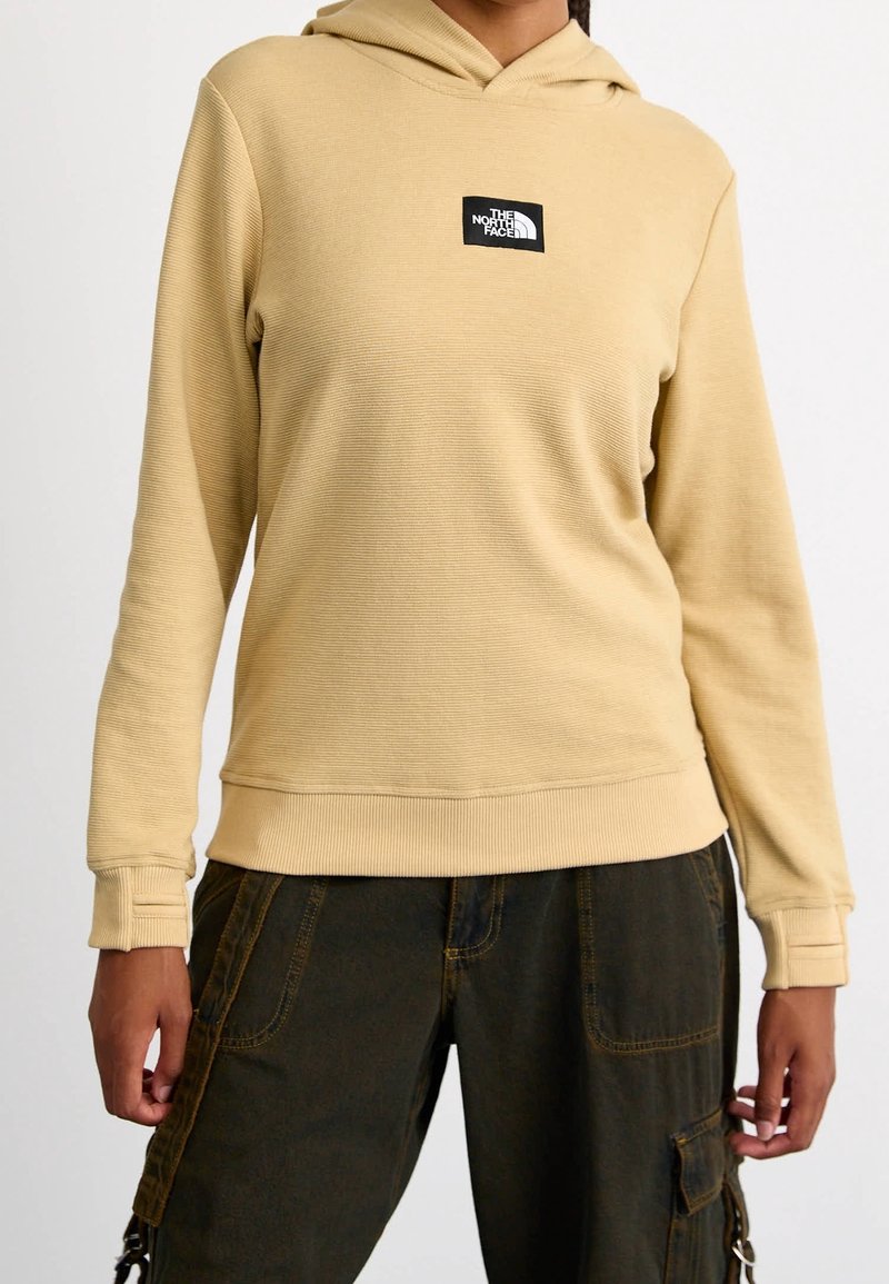 Personne portant un sweat à capuche côtelé beige avec le logo The North Face sur la poitrine et un pantalon cargo noir avec des poches à coutures orange.