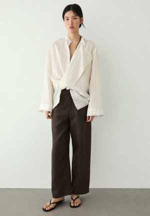 Vrouw staand, gekleed in een losse witte blouse met een gewikkelde textuur, wijd uitlopende donkerbruine broek en zwarte teenslippers tegen een effen achtergrond.