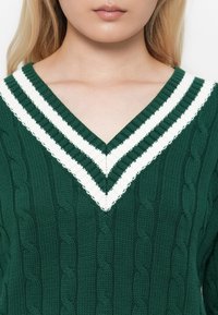 Gros plan sur une personne portant un pull en mailles vertes avec un col en V à double rayures blanches.