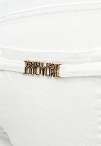 Επίχρυση μεταλλική πλακέτα με το λογότυπο "Versace Jeans Couture" προσαρτημένη στη πίσω τσέπη λευκών τζιν.