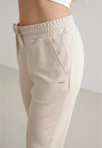 Beige Jogginghose aus weichem Stoff, mit elastischem Bund, Kordelzug, Seitentaschen und einem kleinen metallischen Logoakzent.