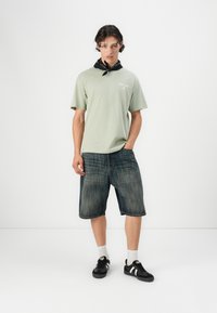 Lys grøn T-shirt med "Palm Coast" tryk, parret med mørke denimshorts. Sorte sneakers med hvide striber og et sort bandana om halsen.
