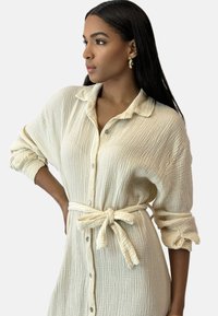 Abito camicia in tessuto cremoso e testurizzato, con collo, maniche arrotolate e cintura annodata in vita. Bottoni silver aggiungono un tocco di dettaglio.