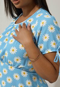 Vaaleansininen lyhythihainen mekko, jossa on valkoisia kamomilloja ja keltaisia keskustoja. Mekossa on kootut kaula-aukot ja nauhat hihansuissa, yhdistettynä kultaketjuun.