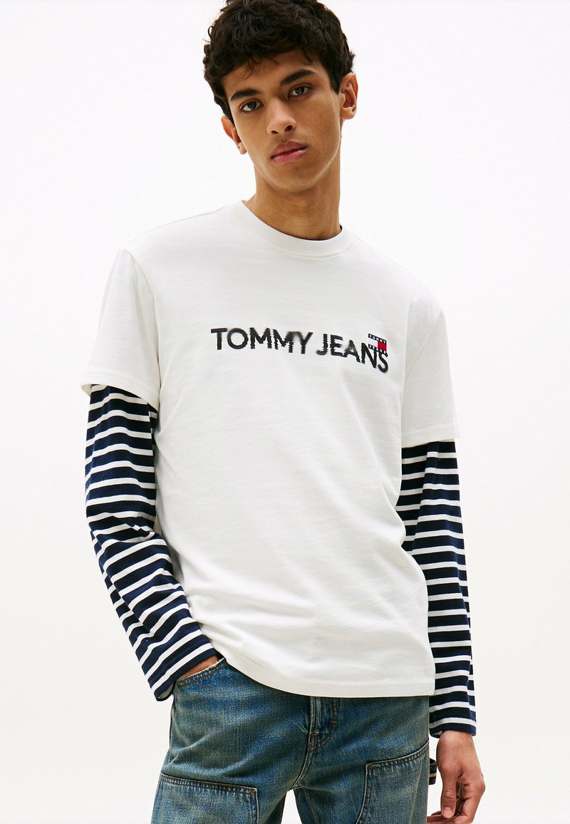 Camiseta blanca con texto negro "TOMMY JEANS". Mangas largas a rayas en azul marino y blanco. Combinada con unos jeans de mezclilla azul. Diseño simple y casual.
