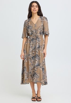 Kvinde står iført en beige midi-kjole med blå blomstrede paisley-mønstre, korte ærmer, V-hals og sorte hælede sandaler med stropper.