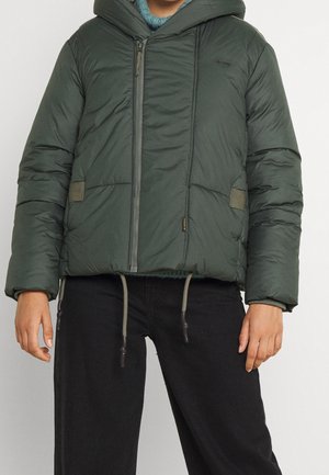 Personne portant une veste matelassée vert foncé avec capuche et fermeture éclair à l'avant, associée à un pantalon noir, debout devant un fond uni.