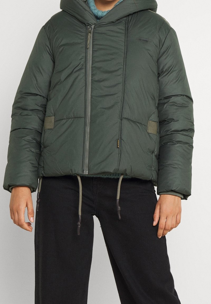 Personne portant une veste matelassée vert foncé avec capuche et fermeture éclair à l'avant, associée à un pantalon noir, debout devant un fond uni.