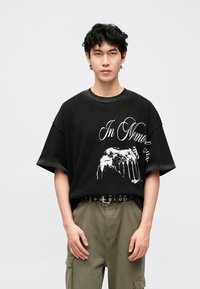 Νέος άνδρας με σγουρά μαύρα μαλλιά που φοράει μαύρο oversized T-shirt με λευκό κείμενο και γραφικό, ελαιδί cargo παντελόνι και μαύρη ζώνη.