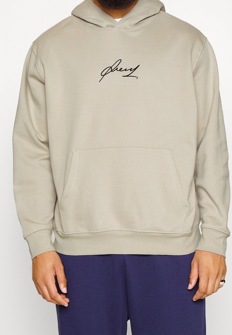 Hoodie beige con tasca a marsupio frontale, dotato di un logo nero in stile firma sul petto e polsini a coste. Materiale morbido e vestibilità comoda.