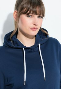 Felpa con cappuccio blu navy, dotata di lacci a contrasto bianchi con punte in metallo. Tessuto morbido, vestibilità comoda, con un cappuccio ampio.