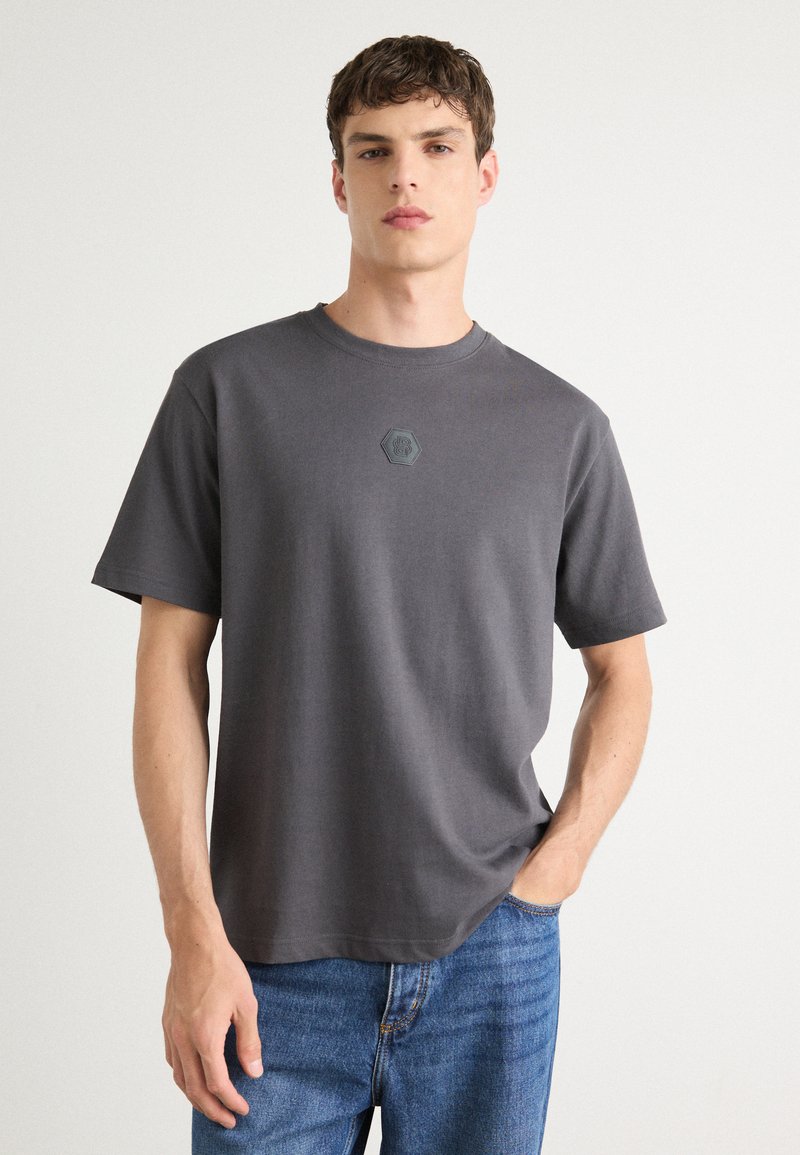 T-shirt en coton gris avec des manches courtes, un col rond et un détail de logo hexagonal au centre. Assorti à un jean en denim bleu.
