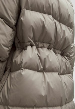 Pufferjack in zacht taupe stof, met een geborduurd ontwerp met kamers, elastische taille en gladde textuur. Geen zichtbare hardware.