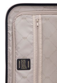 Light beige suitcase interior with embossed logo pattern, zippered compartments, mesh section, and a black tag with care instructions.