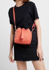 Osoba w czarnym t-shircie i czarnej spódnicy dżinsowej, z koralową torebką na ramię, która ma okrągłe logo i brelok.