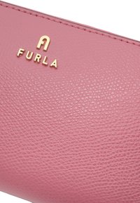 Superficie de cuero texturizado rosa con el logo metálico dorado "FURLA" y un emblema estilizado de "A" bordado a lo largo del borde.