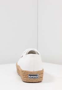 White canvas sneaker with jute rope sole, featuring a visible brand logo. Rounded heel and textured upper with laces.