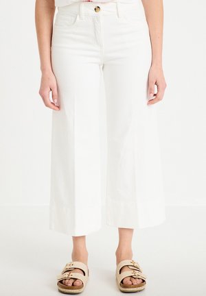 Femme portant un pantalon blanc taille haute à jambes larges, raccourci au-dessus des chevilles, et des sandales tressées beiges avec boucle sur fond blanc.