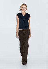 Marineblauw mouwloos topje met een V-hals, gecombineerd met bruine ribfluwelen broeken met een slim fit en een gestructureerde afwerking. Zwarte hangende ketting.