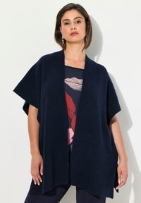 Mörkblå stickad cardigan med vida ärmar och öppen front, som har ett strukturerat ribbmönster. Bärs över en mönstrad topp.