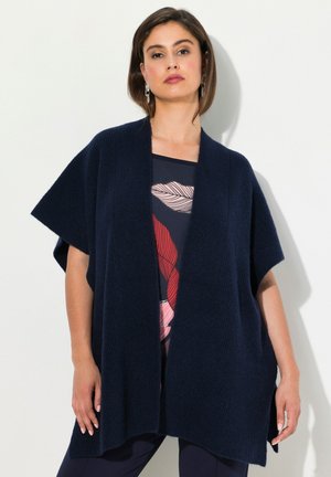 Gilet tricoté bleu marine avec des manches larges et une ouverture sur le devant, présentant un motif en côtes texturé. Porté sur un haut à motifs.