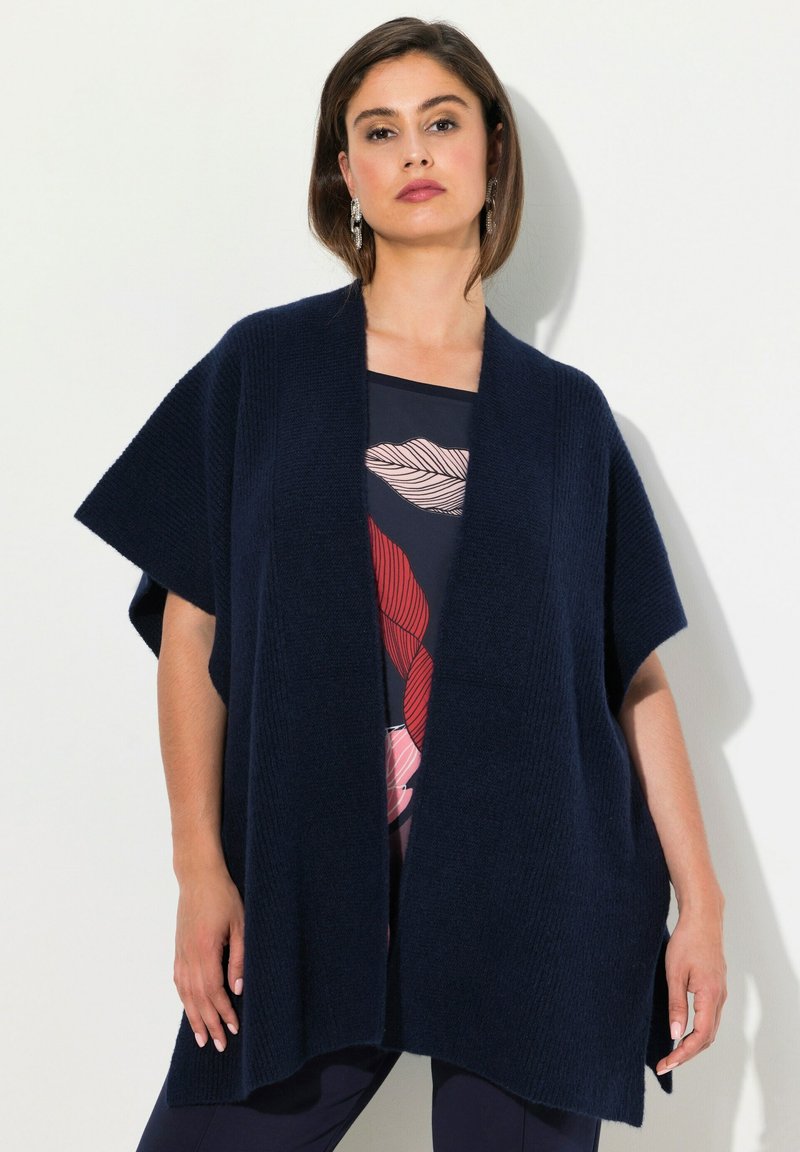 Mörkblå stickad cardigan med vida ärmar och öppen front, som har ett strukturerat ribbmönster. Bärs över en mönstrad topp.