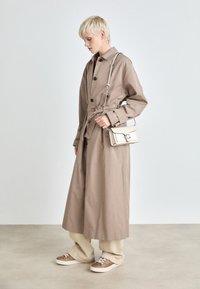 Lång beige trenchcoat med bälte i midjan och svarta knappar, tillsammans med en ljusfärgad crossbody-väska och mönstrade sneakers.
