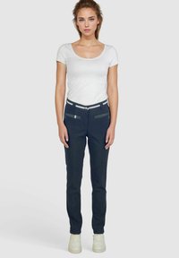 Pantalons noirs ajustés avec une taille à rayures blanches, poches zippées à l'avant et un tissu lisse et texturé. Portés avec un haut blanc ajusté.