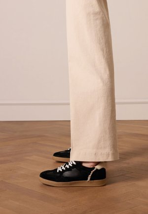 Black suede sneakers with white laces, textured sides, and gum soles. Paired with light beige wide-leg trousers, set against wooden flooring.