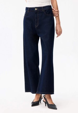 Kobieta ubrana w ciemnoniebieskie dżinsy o szerokich nogawkach i czarne czółenka na niskim obcasie ze zapięciem z tyłu, stojąca na białym tle.