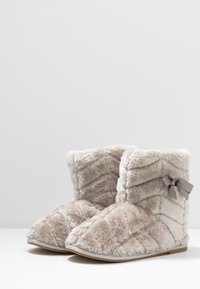 Bottes cheville beige duveteuses avec texture en chevrons, semelle plate, et nœud décoratif gris sur le côté. Matière douce en peluche et design à bout arrondi.
