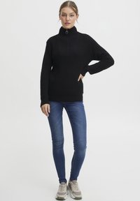Fekete kötött pulóver magas gallérral és cipzár részletekkel, kék skinny farmernadrággal és szürke sportcipőkkel párosítva. Simább textúra és laza fazon.