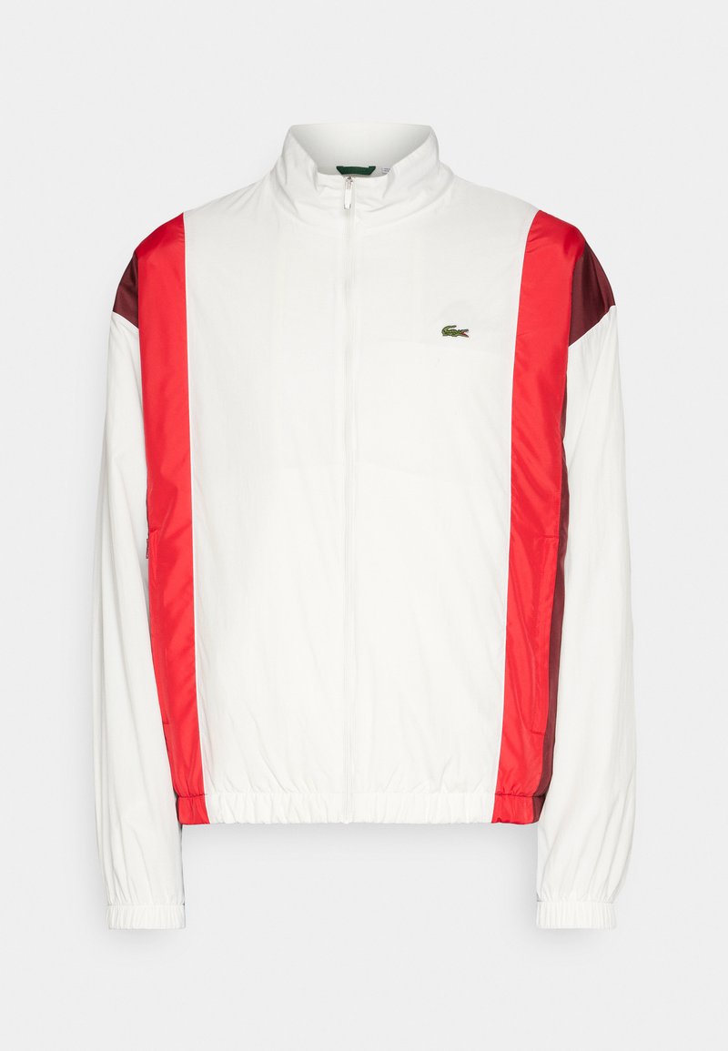 Veste de sport blanche avec des rayures rouges et bordeaux sur les épaules, fermeture éclair, poignets élastiques et logo Lacoste sur la poitrine. Texture lisse.