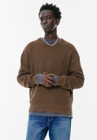 Bruine geribbelde trui met lange mouwen, over een grijze langemouwen shirt heen gedragen. Zilveren ketting voegt detail toe. G gedragen met blauwe jeans.