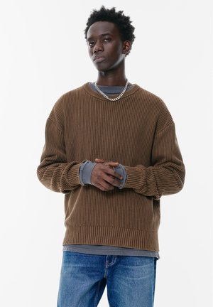 Bruine geribbelde trui met lange mouwen, over een grijze langemouwen shirt heen gedragen. Zilveren ketting voegt detail toe. G gedragen met blauwe jeans.
