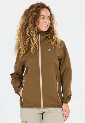 Frau mit lockigem Haar, die eine braune Jacke mit Reißverschluss und Reißverschlusstaschen trägt, steht mit den Händen in den Taschen und lächelt seitlich.