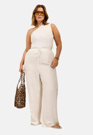 Kobieta ubrana w biały top na jedno ramię i szerokie spodnie, trzyma brązową plecioną torbę, z żółtymi okularami przeciwsłonecznymi i złotą bransoletką.