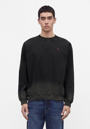 Jeune homme aux cheveux courts et foncés portant un sweat-shirt noir délavé et un jean bleu, debout devant un fond blanc uni.