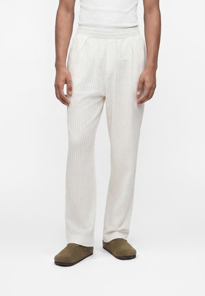 Άνδρας που φοράει λευκό παντελόνι με υφή, λευκό αμάνικο τοπ και πράσινα ελιάς slip-on παπούτσια, στέκεται μπροστά σε απλό λευκό φόντο.