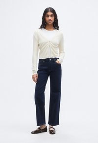 Μπεζ ριπ ζακέτα με κούμπωμα από μπροστά, σετ με τζιν παντελόνι ευρύ πόδι σε σκούρο μπλε με ψηλή μέση. Καφέ παπούτσια με λουράκι διακοσμητικό.