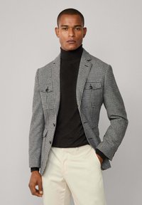 Checkered blazer in black and white with a tailored fit, two front pockets, and a notch lapel, worn over a black turtleneck. Light beige trousers.