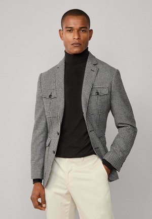 Schwarz-weißer karierten Blazer in taillierter Passform, mit zwei Vorderaschen und Reverskragen, über einem schwarzen Rollkragenpullover getragen. Helle beiger Hose.