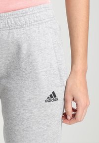 Szare dresy wykonane z mieszanki bawełny, z elastycznym pasem i kieszeniami bocznymi. Logo Adidas jest nadrukowane na czarno na lewej nodze.