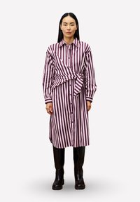 Frau in einem langärmeligen, lila und weißen, vertikal gestreiften Kleid mit geknotetem Taillengürtel, kombiniert mit schwarzen kniehohen Stiefeln, stehend vor einem weißen Hintergrund.